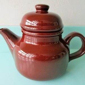 May include: A brown ceramic teapot with a lid. The teapot has a rounded body and a handle on the side.
