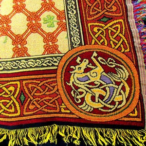 May include: A woven throw blanket featuring a cream-colored section with an orange lattice pattern and green shamrocks, bordered by red and yellow Celtic knotwork. A circular medallion displays a stylized dragon, finished with yellow fringe.