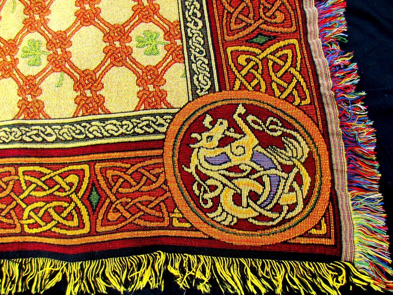 May include: A woven throw blanket featuring a cream-colored section with an orange lattice pattern and green shamrocks, bordered by red and yellow Celtic knotwork. A circular medallion displays a stylized dragon, finished with yellow fringe.