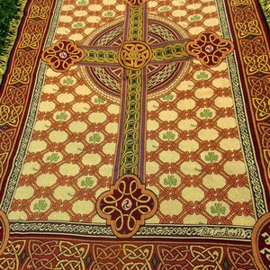 May include: A woven throw blanket featuring a large central Celtic cross in shades of red, gold, and green against a cream background. The design includes a repeating lattice pattern with small green shamrocks and a decorative border with Celtic knots.