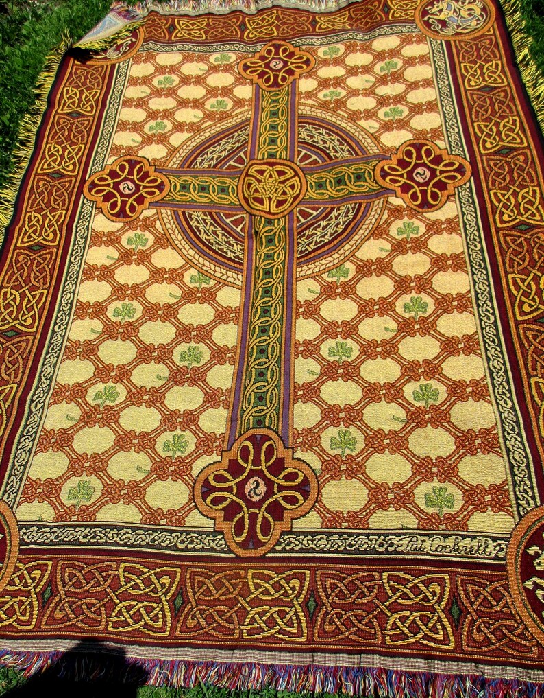 May include: A woven throw blanket featuring a large central Celtic cross in shades of red, gold, and green against a cream background. The design includes a repeating lattice pattern with small green shamrocks and a decorative border with Celtic knots.