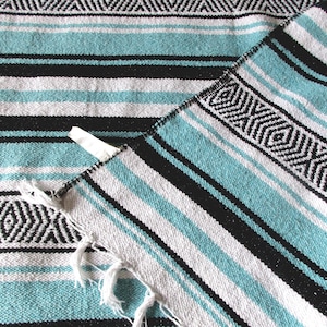 May include: A woven blanket featuring a repeating pattern of horizontal stripes in turquoise, black, and white. The design includes a geometric diamond border motif and a fringed edge with a small white fabric tag attached to the corner seam.
