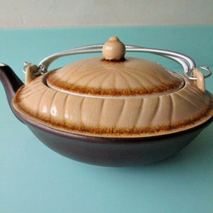 May include: A brown and beige ceramic teapot with a spring-loaded lid and handle. The teapot has a decorative pattern of brown lines and a brown rim.