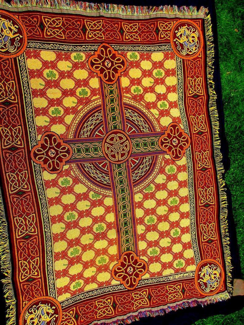 May include: A woven throw blanket featuring a large central Celtic cross design in red, gold, and green. The background has a repeating lattice pattern with small green shamrocks. The border displays intricate knotwork and corner medallions.