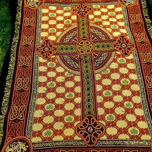 May include: A woven throw blanket featuring a large central Celtic cross design on a cream background with a red lattice pattern and green shamrocks. The border displays intricate knotwork and corner medallions with horse motifs and fringe.