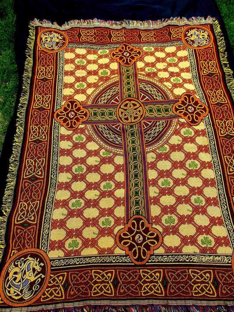 May include: A woven throw blanket featuring a large central Celtic cross design on a cream background with a red lattice pattern and green shamrocks. The border displays intricate knotwork and corner medallions with horse motifs and fringe.
