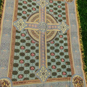 May include: A woven throw blanket featuring a large central Celtic cross design in yellow and blue, set against a dark green background with a repeating pattern of small pink shamrocks. The border displays intricate knotwork and corner dragon motifs.