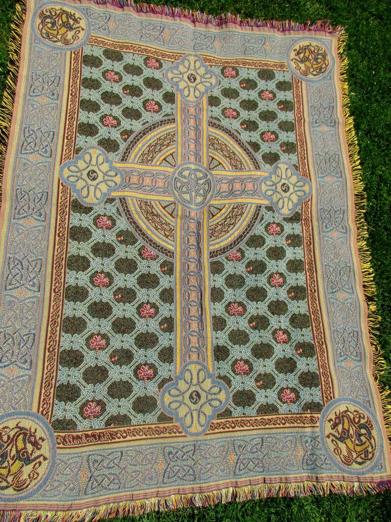 May include: A woven throw blanket featuring a large central Celtic cross design in yellow and blue, set against a dark green background with a repeating pattern of small pink shamrocks. The border displays intricate knotwork and corner dragon motifs.