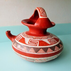 May include: A red clay pot with a handle and a spout. The pot is decorated with white and brown geometric patterns.