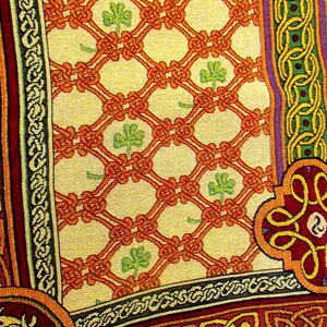 May include: A woven textile featuring a cream-colored background with an orange lattice pattern and small green shamrocks. The border displays intricate knotwork in shades of red, green, and yellow, with a stylized animal figure in the corner.