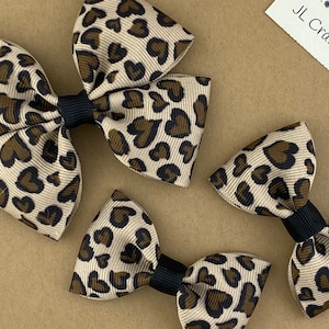May include: Three brown and black leopard print hair bows with black centers. The bows are different sizes. The largest bow is a double bow. The smaller bows are single bows. The bows are on a brown background.