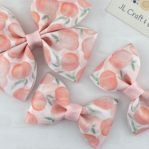 May include: Three peach-themed hair bows in different sizes. The bows are made of white fabric with a peach print and have pink centers.
