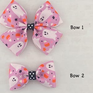 May include: Two pink and white hair bows with a ghost and candy corn pattern. The larger bow is labeled "Bow 1" and the smaller bow is labeled "Bow 2".