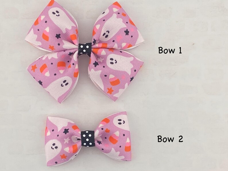 May include: Two pink and white hair bows with a ghost and candy corn pattern. The larger bow is labeled "Bow 1" and the smaller bow is labeled "Bow 2".