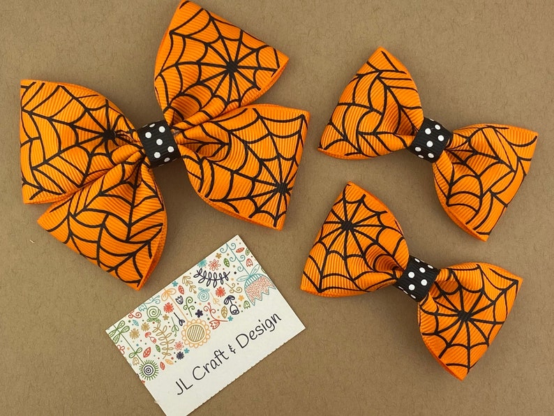 Puede incluir: Tres lazos para el pelo de color naranja y negro con estampado de telara&ntilde;a y centros de lunares negros y blancos. Los lazos est&aacute;n dispuestos en orden decreciente de tama&ntilde;o de izquierda a derecha.