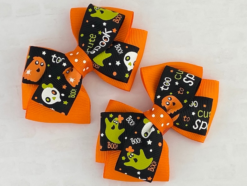 May include: Two orange and black Halloween hair bows with white polka dots. The bows feature a black ribbon with green ghosts, stars, and the text "too cute to spook" and "too cute to spook".