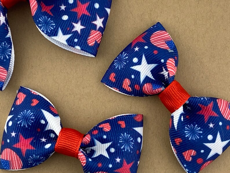 May include: Three red, white, and blue bow ties with a pattern of stars, hearts, and fireworks. The bow ties are made of fabric and have a red center.