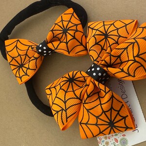 Puede incluir: Dos lazos para el pelo de color naranja y negro con estampado de telara&ntilde;a y centros de lunares negros y blancos unidos a una diadema el&aacute;stica negra.