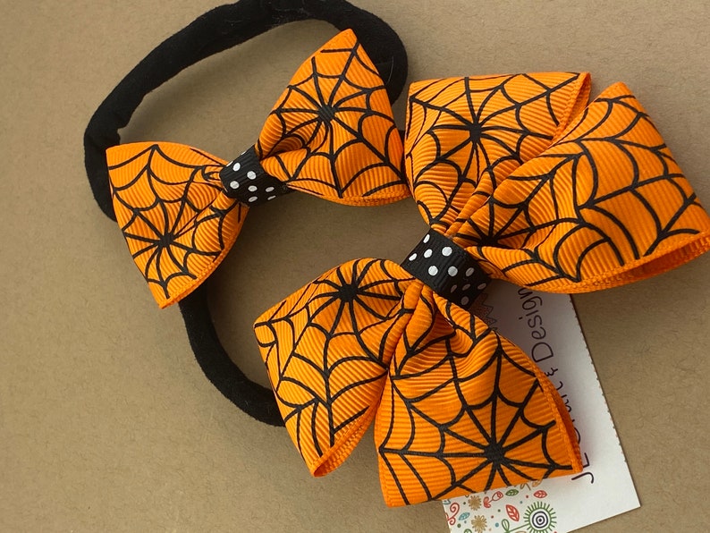 Puede incluir: Dos lazos para el pelo de color naranja y negro con estampado de telara&ntilde;a y centros de lunares negros y blancos unidos a una diadema el&aacute;stica negra.