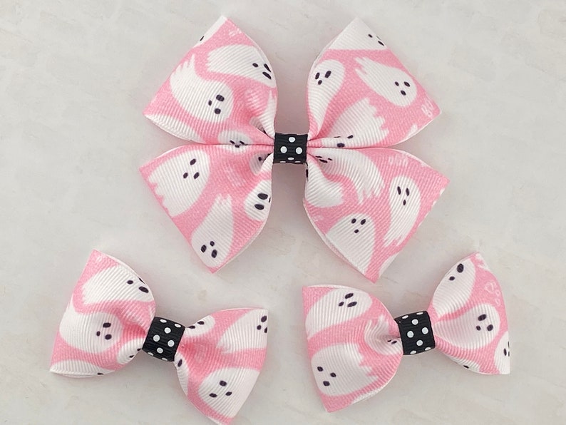 May include: Three pink hair bows with a white ghost pattern. The largest bow is a double bow with black polka dot accents. The two smaller bows are single bows with black polka dot accents.