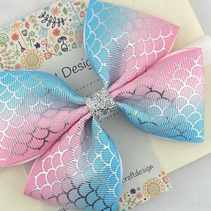 May include: A pink and blue ombre bow with a mermaid scale pattern and silver glitter in the center. The bow is attached to a white elastic headband.