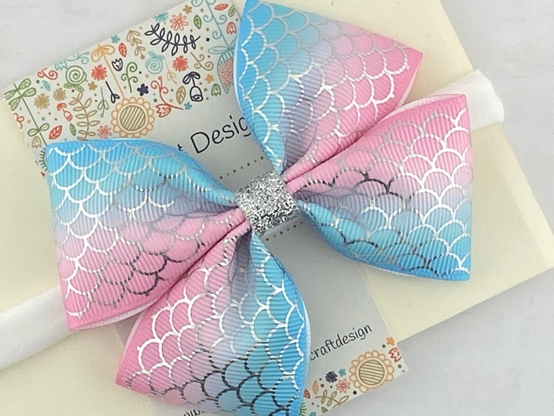 May include: A pink and blue ombre bow with a mermaid scale pattern and silver glitter in the center. The bow is attached to a white elastic headband.