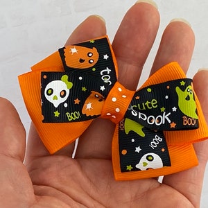 May include: Orange and black Halloween hair bow with a ghost, skull, and stars pattern. The bow has the text "too cute spook" and "boo!" printed on it.