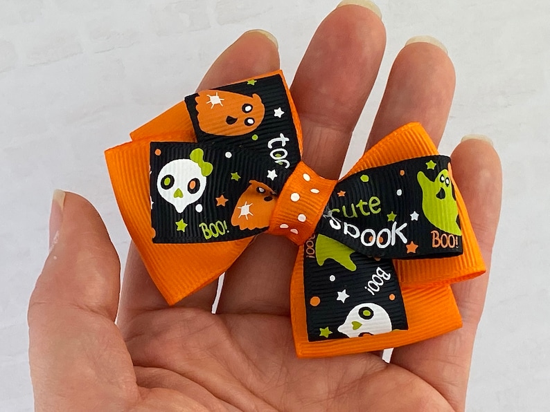 May include: Orange and black Halloween hair bow with a ghost, skull, and stars pattern. The bow has the text "too cute spook" and "boo!" printed on it.