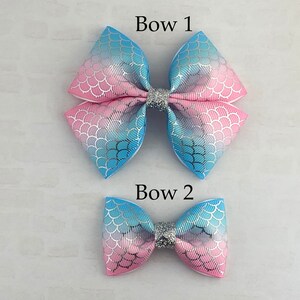 May include: Two mermaid-themed hair bows, one larger than the other. Both bows are pink and blue with a silver glitter center. The larger bow is labeled "Bow 1" and the smaller bow is labeled "Bow 2".