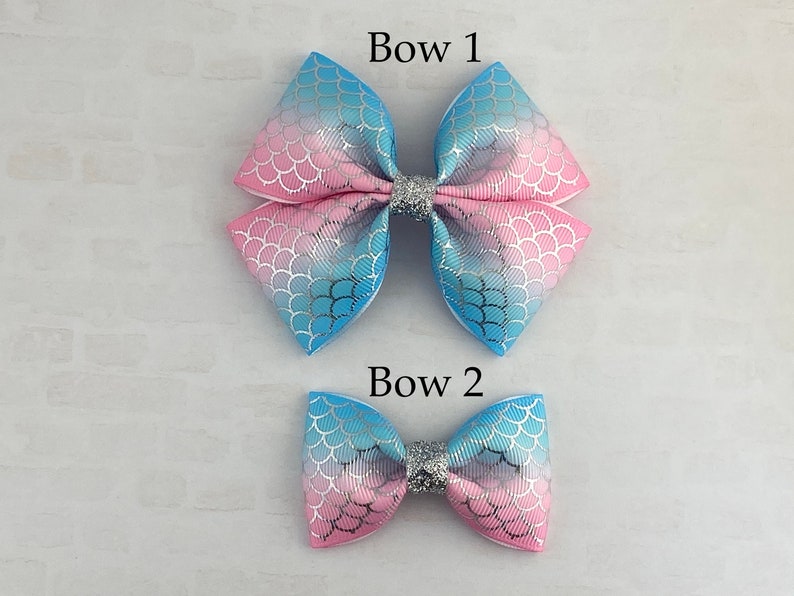 May include: Two mermaid-themed hair bows, one larger than the other. Both bows are pink and blue with a silver glitter center. The larger bow is labeled "Bow 1" and the smaller bow is labeled "Bow 2".