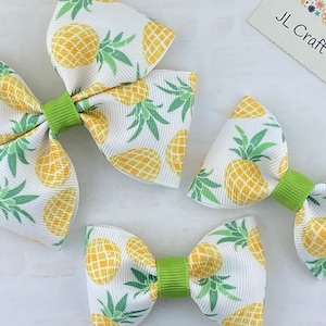May include: Three pineapple-themed hair bows in different sizes. The largest bow is white with green leaves and yellow pineapples. The medium bow is white with green leaves and yellow pineapples. The smallest bow is white with green leaves and yellow pineapples. All bows have a green center.