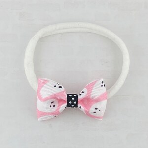 May include: A pink and white patterned bow with black polka dots is attached to a white elastic headband.
