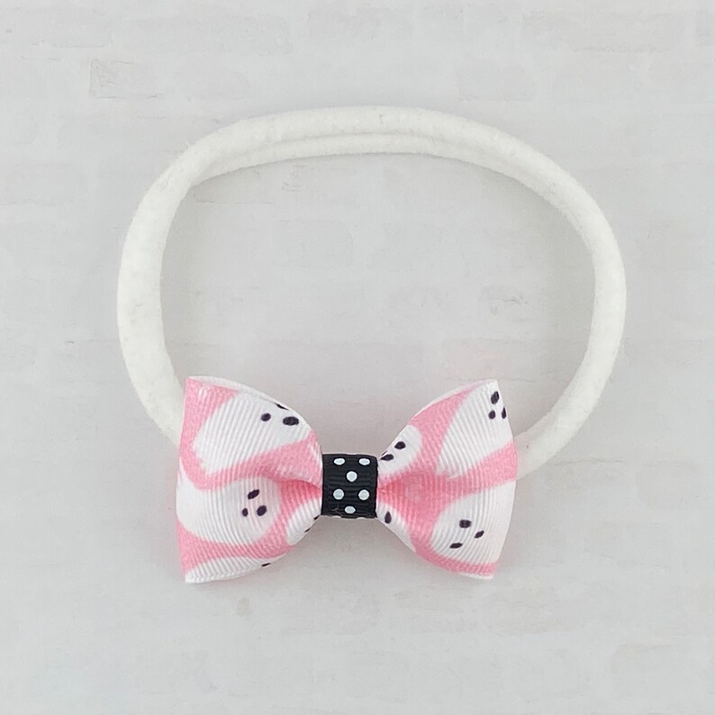 May include: A pink and white patterned bow with black polka dots is attached to a white elastic headband.