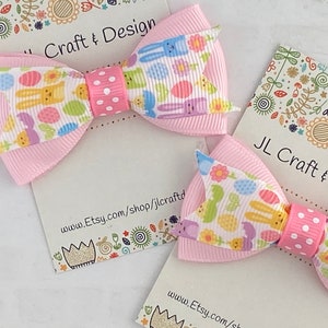 Pu&ograve; includere: Due fiocchi per capelli rosa con uno sfondo bianco con un motivo di coniglietti di Pasqua, uova e fiori. I fiocchi sono legati con un nastro a pois bianchi. I fiocchi sono esposti su una carta bianca con il testo "JL Craft & Design" e un indirizzo di sito web.