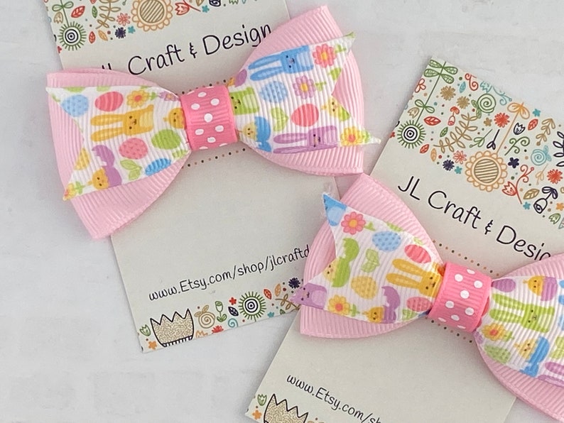 Pu&ograve; includere: Due fiocchi per capelli rosa con uno sfondo bianco con un motivo di coniglietti di Pasqua, uova e fiori. I fiocchi sono legati con un nastro a pois bianchi. I fiocchi sono esposti su una carta bianca con il testo "JL Craft & Design" e un indirizzo di sito web.