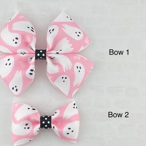 May include: Two pink hair bows with a white ghost pattern and black polka dot accents. The larger bow is labeled "Bow 1" and the smaller bow is labeled "Bow 2".