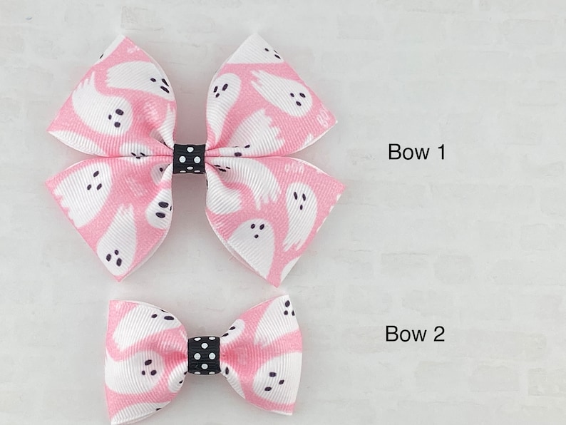 May include: Two pink hair bows with a white ghost pattern and black polka dot accents. The larger bow is labeled "Bow 1" and the smaller bow is labeled "Bow 2".