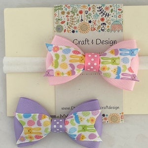 Pu&ograve; includere: Due fiocchi per capelli con un elastico bianco per capelli. I fiocchi sono realizzati in nastro rosa e viola con una stampa bianca e pastello di coniglietti e uova. Il testo "Craft & Design" &egrave; stampato sulla carta.