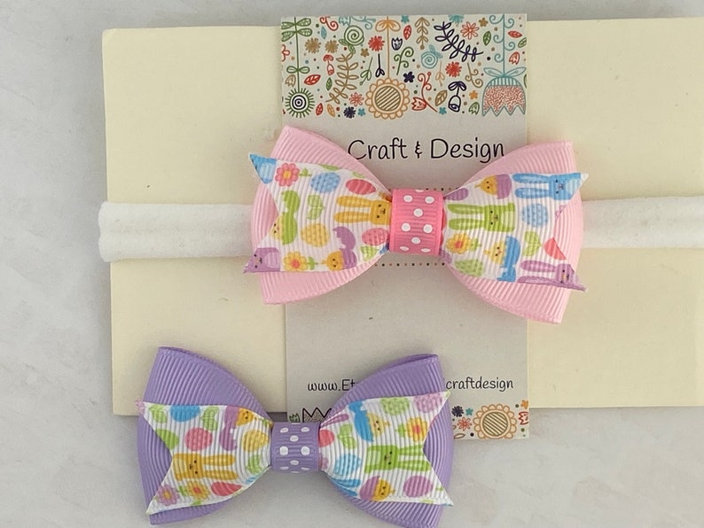 Pu&ograve; includere: Due fiocchi per capelli con un elastico bianco per capelli. I fiocchi sono realizzati in nastro rosa e viola con una stampa bianca e pastello di coniglietti e uova. Il testo "Craft & Design" &egrave; stampato sulla carta.