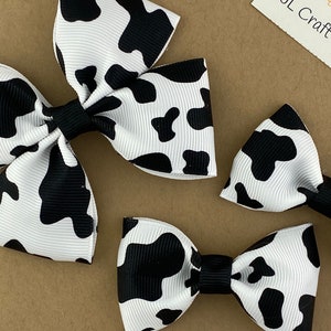 May include: Three cow print hair bows in different sizes. The largest bow is a white bow with black cow print spots. The medium bow is a black bow with white cow print spots. The smallest bow is a white bow with black cow print spots.