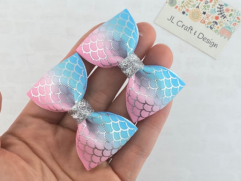 May include: Two pink and blue ombre hair bows with a mermaid scale pattern and silver glitter accents. JL Craft & Design.
