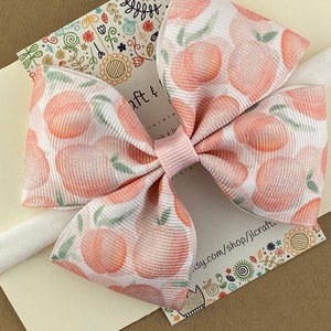 May include: A white headband with a pink bow and a peach print. The bow is made of a soft, textured fabric and has a white center. The headband is made of a soft, stretchy material.