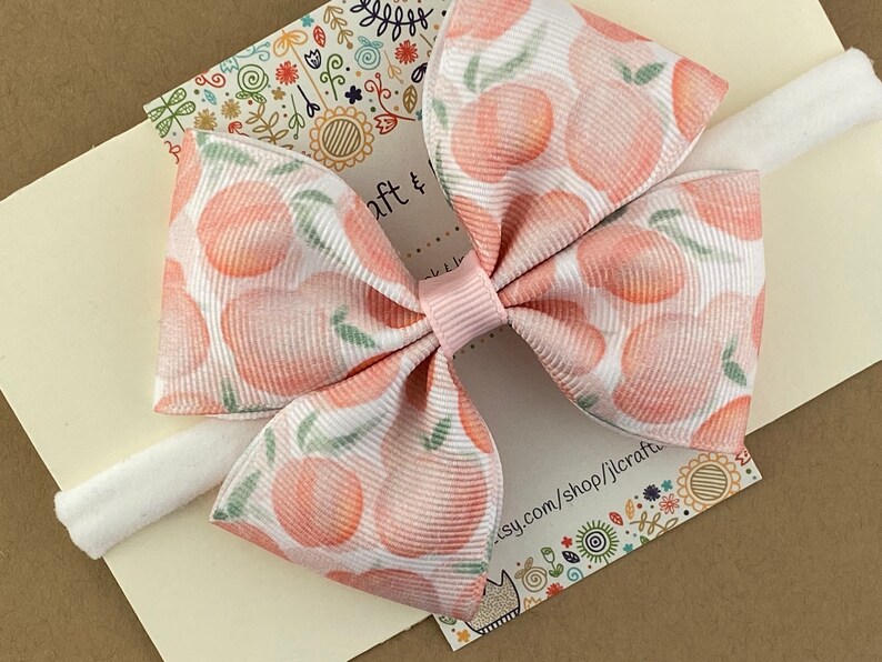 May include: A white headband with a pink bow and a peach print. The bow is made of a soft, textured fabric and has a white center. The headband is made of a soft, stretchy material.