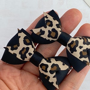 Peut inclure: Deux petits n&oelig;uds pour cheveux avec un imprim&eacute; l&eacute;opard noir et beige. Les n&oelig;uds sont faits de ruban gros-grain, avec une base noire et un motif l&eacute;opard beige. Les n&oelig;uds sont tenus dans la main d'une personne.