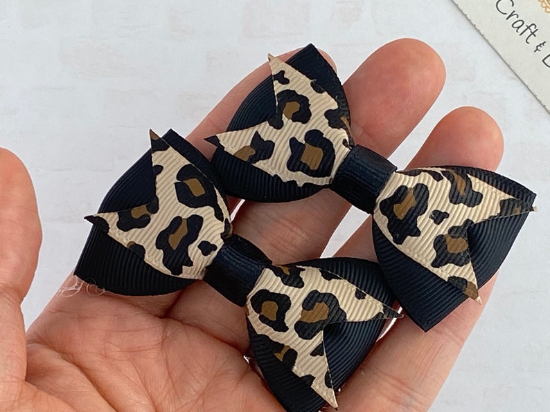 Peut inclure: Deux petits n&oelig;uds pour cheveux avec un imprim&eacute; l&eacute;opard noir et beige. Les n&oelig;uds sont faits de ruban gros-grain, avec une base noire et un motif l&eacute;opard beige. Les n&oelig;uds sont tenus dans la main d'une personne.