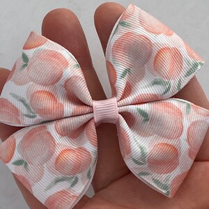 May include: A white and pink hair bow with a peach print. The bow is made of a soft, textured fabric and has a pink center.