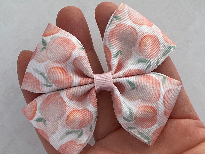May include: A white and pink hair bow with a peach print. The bow is made of a soft, textured fabric and has a pink center.
