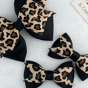 May include: Several black hair bows with a tan and brown leopard print pattern. The bows are made of a grosgrain ribbon. The image also includes a small card with the text "JL Craft & Design".