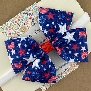 May include: A red, white, and blue star and heart patterned hair bow on a white elastic headband.