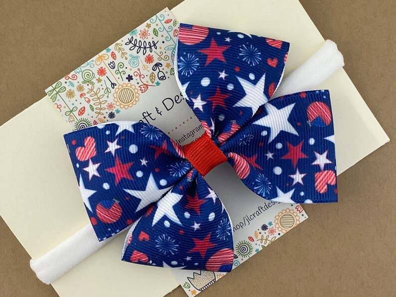 May include: A red, white, and blue star and heart patterned hair bow on a white elastic headband.